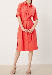 Vestito camicia corallo con maniche corte, cintura in vita, chiusura frontale a bottoni, spacchi laterali e una texture di tessuto morbido. Indossato con sandali beige senza lacci.