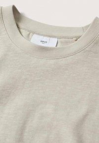 Gros plan d'un t-shirt beige à col rond avec une étiquette blanche visible indiquant la marque "MNG MAN" à l'intérieur du col.