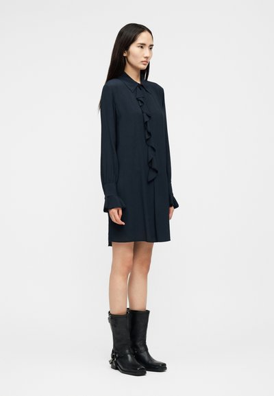 Γυναίκα στέκεται φορώντας μαύρο φόρεμα με μακριά μανίκια και βολάν στο μπροστινό μέρος, μαζί με μαύρες δερμάτινες μπότες μέχρι τη μέση της γάμπας, σε λευκό φόντο.