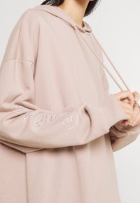 Helle rosa Kapuzenjacke aus weichem Material, mit lockerer Passform, Kordelzugkapuze und dekorativen Strassakzenten an den Ärmeln.