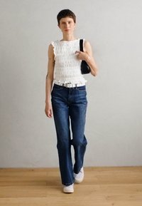 White, textured sleeveless top with ruffle trim; high-waisted dark blue jeans; white sneakers; small black bag; wooden floor backdrop.