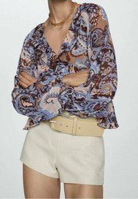 Blouse à manches longues avec un motif floral marron, bleu et rose, col froufrouté, associée à un short beige et une ceinture assortie.