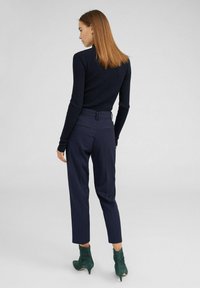 Marineblaues, geripptes Langarmoberteil, kombiniert mit maßgeschneiderten marineblauen Hosen. Das Modell trägt grüne Stiefeletten mit einem dezenten Absatz. Schlichtes, elegantes Design.