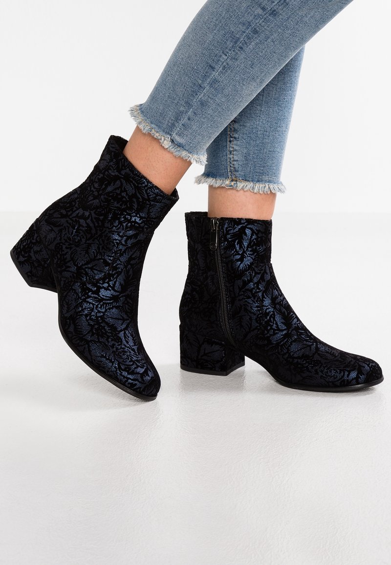 Bottines en velours noir avec motif floral et fermeture éclair latérale portées avec un jean bleu à ourlet effiloché.