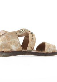 Sandale ouverte en suède beige usé avec des clous en laiton sur des brides croisées et une semelle sombre, vue de côté sur fond blanc.