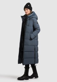 Lang, fyldig mørkeblå frakke med hætte og knaplukning. Har sidelommer og tekstureret stof. Båret med sorte støvler og en hue.