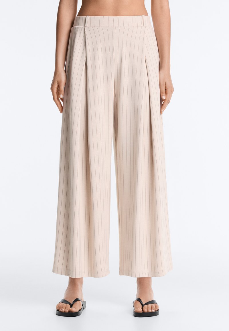 Pantalon beige à jambes larges avec fines rayures verticales, doté de plis et d'une taille haute, porté avec des sandales noires ouvertes à l'avant sur fond clair.