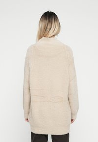 Zachte, beige gebreide cardigan met een hoge kraag en verlaagde schouders. Bevat een strikdetail op de rug en heeft een rechte, comfortabele pasvorm.