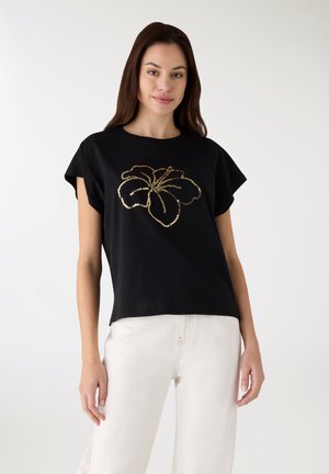 T-shirt nera in cotone con maniche corte e motivo floreale di paillettes dorate sul davanti. Abbinata a pantaloni di colore chiaro.