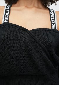 Kvinna som bär en svart stickad topp med synliga svarta och vita märkesband med texten "KARL LAGERFELD JEANS" över axlarna.