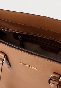 Μεγάλη καφέ δερμάτινη τσάντα ώμου με χρυσαφί εξαρτήματα, που διαθέτει εσωτερική τσέπη με φερμουάρ και λεπτομέρειες με λογότυπο, λεία υφή και δομημένο σχεδιασμό.