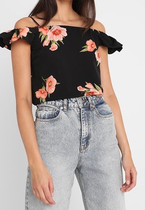 Femme portant un haut noir à épaules dénudées avec des imprimés floraux roses et un jean denim bleu clair taille haute, debout devant un fond uni.
