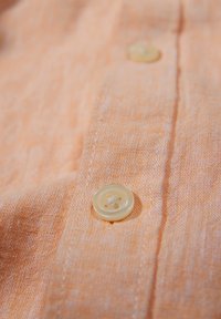 Helles orangefarbiges Gewebe mit strukturierter Oberfläche und einem runden, cremefarbenen Knopf mit vier Löchern und leichtem Glanz.