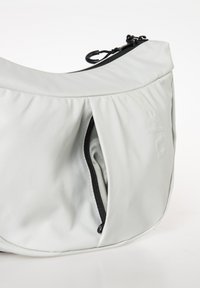 Hellegraue Tasche aus weichem Stoff, mit geschwungener Form, schwarzem Reißverschluss und einer Außentasche. Verfügt über ein dezentes, strukturiertes Logodetail.
