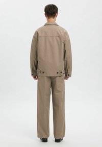 Beige bomuldsjakke med krave og knappedetaljer, kombineret med løstsiddende beige bukser, set bagfra mod en ensfarvet baggrund.