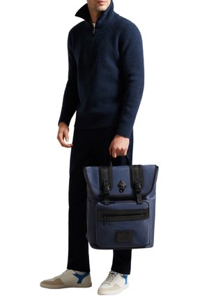 Uomo in maglione blu scuro e pantaloni neri, che tiene uno zaino blu e nero per il manico, indossando sneakers bianche con dettagli blu.
