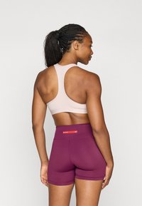 Svetlo roza športni modrček z obliko racerback, v kombinaciji z bordo visokimi hlačami, ki imajo na pasu rdeč logotip Nike.