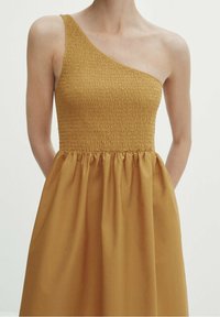 Robe jaune moutarde avec un design asymétrique à une épaule, comprenant un corsage smocké et une jupe volumineuse froncée en tissu tissé lisse.