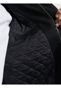 Intérieur de la veste noire avec doublure matelassée à motif en losange et fermeture éclair. Le matériau semble lisse avec un léger brillant.