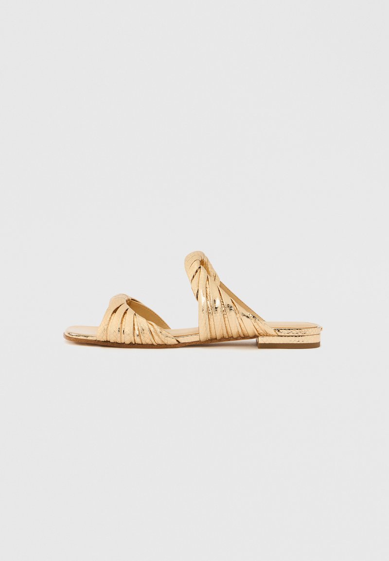 Χρυσά σανδάλια με επίπεδη σόλα και στριφτές λουρίδες, με ορθογώνιο σχέδιο στη μύτη. Το επάνω μέρος έχει ελαφρώς γυαλιστερή υφή, με επίπεδη σόλα.