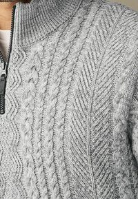 Szary sweter dzianinowy z zamkiem błyskawicznym, z teksturowanymi wzorami w warkocze, ściągaczem przy kołnierzu oraz mieszaniem wypukłych i płaskich ściegów.