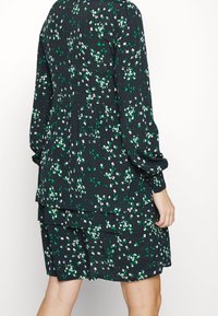 Vestido oscuro de flores con hojas verdes y pequeñas flores blancas, de mangas largas y un diseño en capas y acampanado. Tela suave y texturizada.