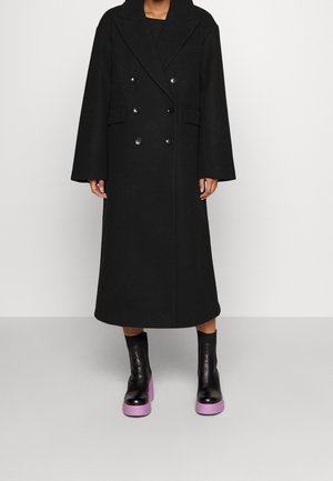 Personne portant un long manteau noir croisé à larges revers et des bottines noires avec des semelles épaisses violettes.