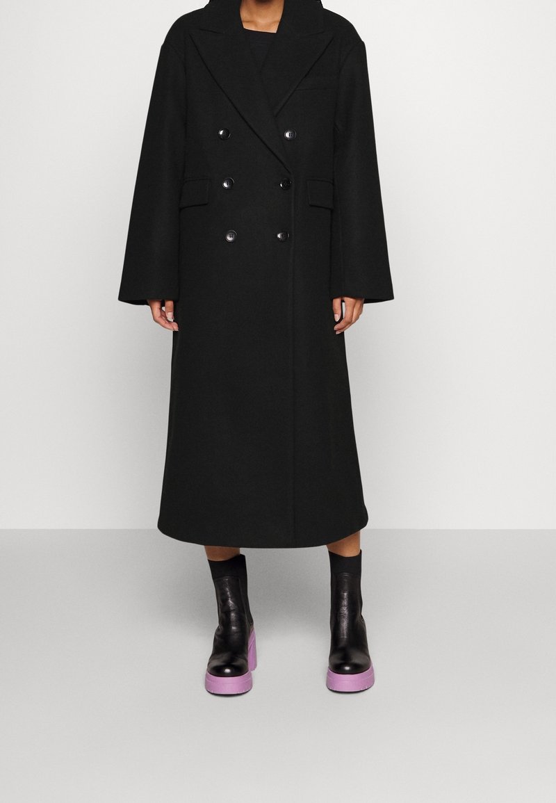 Personne portant un long manteau noir croisé à larges revers et des bottines noires avec des semelles épaisses violettes.