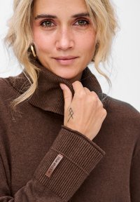 Kobieta z blond włosami, ubrana w brązowy sweter z golfem, trzymająca kołnierz lewą ręką, na której widoczny jest geometryczny tatuaż.