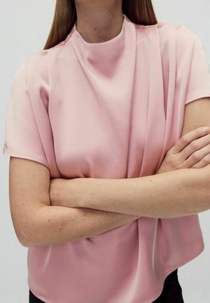 Personne portant un chemisier à manches courtes rose pâle avec un drapé subtil, les bras croisés, sur un fond clair uni.