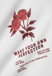 Balts audums ar sarkanu grafiku, kurā redzami divi ziedi un teksts: "VEIDO SAVU IDEĀLU" ar papildu frāzēm zemāk biezā fontā.