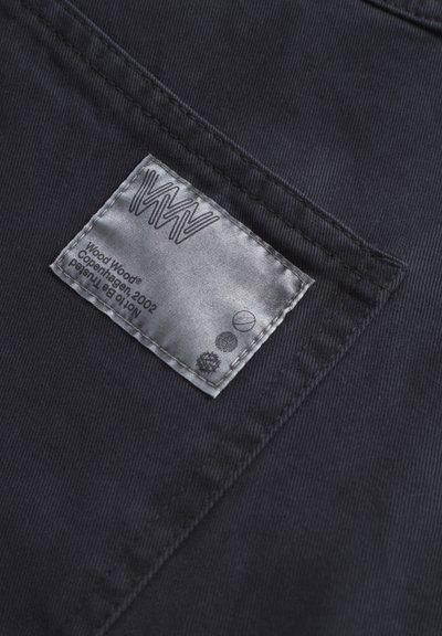 Material textil negru cu o textură în dungi, având o etichetă de întreținere argintie cu inscriptionarea "Wood Wood" și simboluri. Eticheta este poziționată pe cusătura unei buzunare din spate.