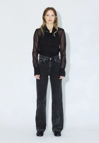 Camicia nera trasparente con bottoni e maniche lunghe, abbinata a jeans neri a vita alta e svasati. Il modello è in piedi contro uno sfondo grigio chiaro.