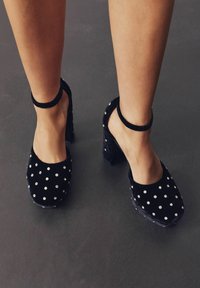 Kojos vilkintis juodus aksominius aukštakulnius batus su kulkšnies dirželiais ir sidabro spalvos kaštonais tamsiai pilkame grindinyje.