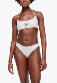 Bikini deux pièces de couleur beige clair, orné d'un motif d'œil en perles sur le haut, avec un bas taille basse assorti.