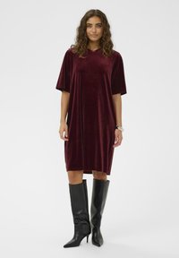 Rochie din catifea bordeaux cu mâneci scurte, croială lejeră, până la genunchi și decolteu rotund. Asortată cu cizme negre cu toc stiletto, până la genunchi.
