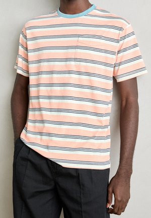 Homme portant un t-shirt à manches courtes rayé pêche, blanc et noir avec une poche sur la poitrine et un pantalon noir, sur un fond uni.