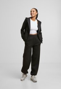 Un sweat à capuche noir avec une fermeture éclair à l'avant, associé à un pantalon de survêtement noir à jambe large assorti. Porté par-dessus un débardeur blanc court ; dispose de poches.