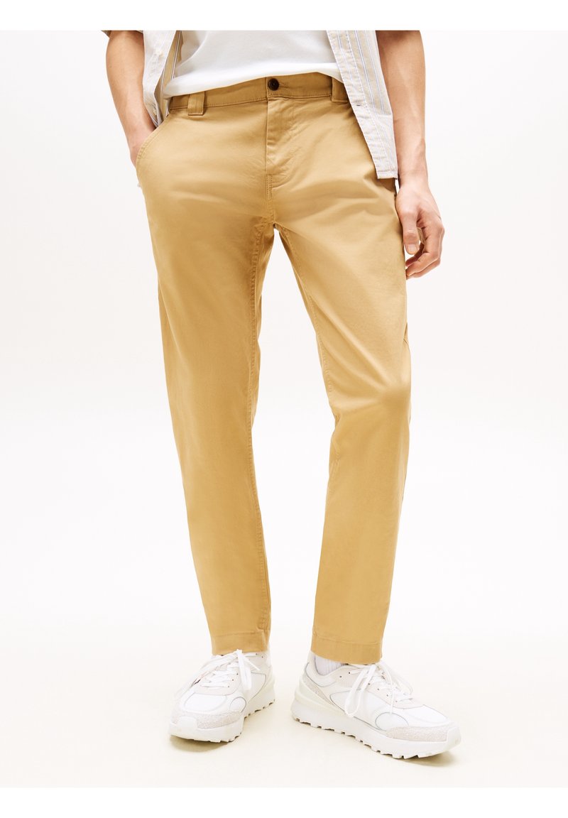 Homme portant un chino beige, des baskets blanches, des chaussettes blanches, un t-shirt blanc et une chemise à rayures claires partiellement visible, debout les mains dans les poches.