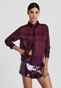 Frau, die eine durchsichtige blütenlilafarbene gestreifte Bluse mit langen Ärmeln und glänzenden blütenlilafarbenen Pailletten-Shorts trägt, steht mit einer Hand auf der Hüfte vor einem schlichten Hintergrund.