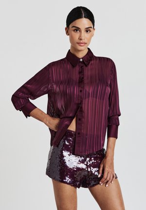 Femme portant un blouse longue à manches transparentes à rayures prune et un short à sequins prune brillant, se tenant avec une main sur la hanche devant un fond uni.