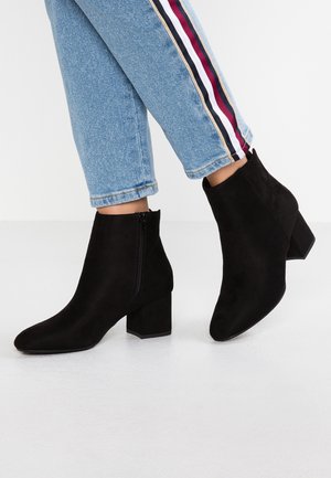 Person trägt hellblaue Jeans mit roten, weißen und schwarzen Seitestreifen sowie schwarze Wildleder-Stiefeletten mit Blockabsätzen und seitlichen Reißverschlüssen.