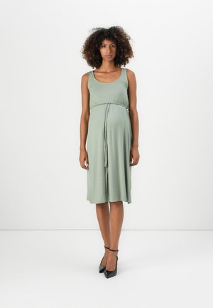 Woman with curly hair standing, wearing a sleeveless sage green midi dress with a tied waist and black ankle-strap heels against a white background.
