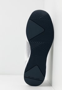 Semelle d'une basket blanche avec une bande de roulement texturée noire affichant des motifs géométriques et la marque "MICHAEL KORS" sur le talon.