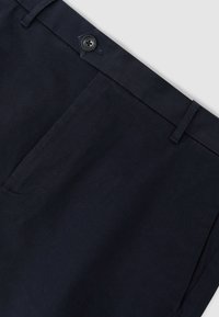 Détail d'un pantalon bleu marine foncé montrant la ceinture avec un bouton noir, une passants de ceinture et une couture de poche latérale.