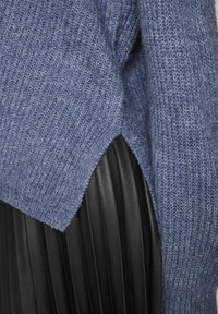 Suéter azul de punto con un patrón texturizado, que cuenta con una abertura lateral que revela una falda de piel sintética con pliegues negra por debajo.