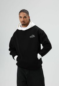 Sweat à capuche noir avec doublure blanche et l'inscription brodée "Boys Champions" sur le devant ; doté d'une poche kangourou et de poignets côtelés.