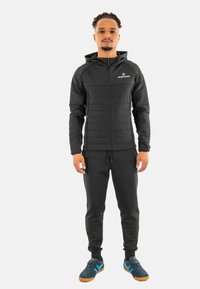 Giacca nera con cappuccio, pannelli frontali trapuntati, chiusura con zip e tasche laterali abbinata a pantaloni scuri e sneakers con dettagli blu.
