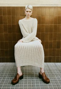 Beige, gestricktes langes Kleid mit weit geschnittenen Ärmeln und offenem Ausschnitt; Modell trägt braune Plateau-Clogs; sitzt an einer braun gefliesten Wand.