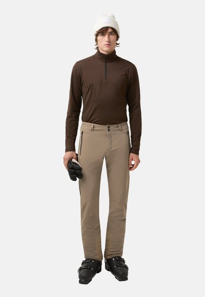 Hnědá dlouhá zipová mikina s vysokým límcem, béžové kalhoty se dvěma zipovými kapsami a černé lyžařské boty. Model drží černé rukavice.